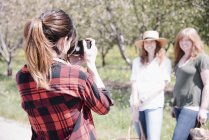 Vista posteriore della fotografa femminile che scatta foto di donne nel frutteto in estate . — Foto stock