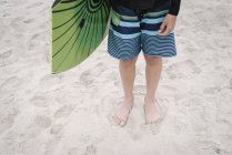 Cropped view of boy standing on sandy beach and holding bodyboard. — Stock Photo