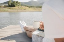 Homem sentado no molhe e segurando caneca e livro no lago . — Fotografia de Stock