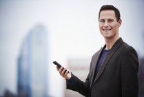 Young businessman standing on rooftop, holding smartphone and looking in camera. — Stock Photo