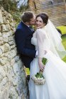 Groom embracing and kissing bride in wedding dress by stone wall outdoors. — Stock Photo