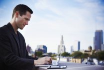 Hombre joven de pie en la azotea del edificio y trabajando con el ordenador portátil con teléfono inteligente al lado . - foto de stock