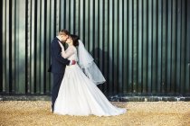 Bride and bridegroom kissing in front of green corrugated metal wall, side view. — Stock Photo
