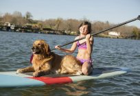 Pre-adolescent girl in swimwear with golden retriever dog on paddleboard on water. — Stock Photo