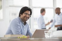 Mature man sitting at desk and using laptop in office with coworkers in background. — Stock Photo