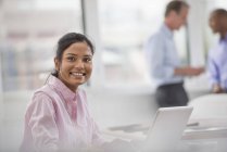 Young woman sitting at desk and using laptop in office with coworkers in background. — Stock Photo