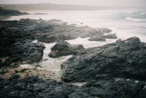 Rocas en la playa por mar con olas rompiendo y la niebla subiendo . - foto de stock
