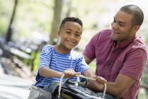 Father helping boy riding old fashioned toy peddle car in sunny park. — Stock Photo