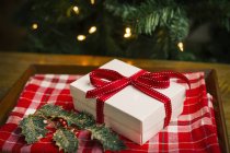 Gift box tied with red ribbon and holly leaves on tablecloth with checker pattern. — Stock Photo