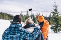 Três pessoas em equipamento de esqui posando para selfie com vara de selfie em montanhas nevadas . — Fotografia de Stock
