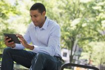 African american man using digital tablet while sitting on park bench. — Stock Photo