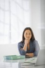 Жінка з рукою на підборідді розслабляється за офісним столом з ноутбуком і зеленою текою . — стокове фото