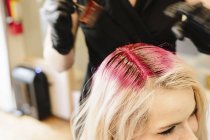 Feminino colorista de cabelo em luvas aplicando tintura de cabelo vermelho para cabelo loiro cliente com escova . — Fotografia de Stock