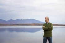Mature man with arms folded by water of lake in mountains. — Stock Photo