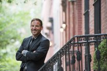Man in suit standing on steps of brownstone building with arms folded. — Stock Photo