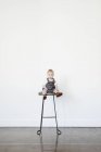 Kleinkind Mädchen im grauen Kleid sitzt auf einem hohen Hocker und blickt in die Kamera. — Stockfoto