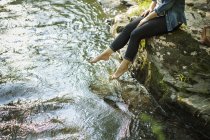 Frau sitzt am Wasserrand und paddelt mit Füßen im Bach. — Stockfoto