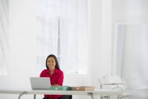 Mediados de adulto mujer asiática sentada en el escritorio con el ordenador portátil y sonriendo en la oficina
. - foto de stock