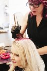 Female hair colorist in gloves applying red hair dye to client blonde hair with brush. — Stock Photo