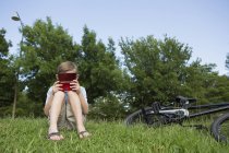 Хлопчик початкового віку сидить на траві на велосипеді і грає в портативну електронну гру . — стокове фото