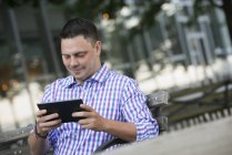 Чоловік середнього віку сидить на лавці і використовує цифровий планшет . — стокове фото