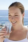 Mujer joven bebiendo vaso de agua clara por mar . - foto de stock