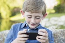 Menino idade elementar ao ar livre inclinando-se contra a rocha e usando jogo eletrônico portátil . — Fotografia de Stock