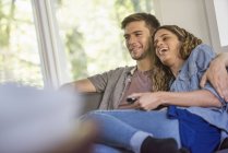 Couple with remote controller sitting side by side laughing and watching tv indoors. — Stock Photo
