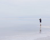 Человек, стоящий на краю затопленных солончаков Бонневилля и фотографирующий в сумерках, Юта, США . — стоковое фото