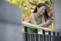 Mujer en vestido verde usando teléfono inteligente al aire libre en el parque de la ciudad bajo los árboles en flor . - foto de stock