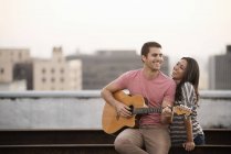 Homem tocando guitarra para mulher no terraço com vista para a cidade ao entardecer . — Fotografia de Stock