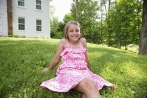 Pre-adolescent girl in pink sundress sitting on lawn in farmhouse garden. — Stock Photo