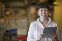 Asian woman holding digital tablet and looking in camera indoors. — Stock Photo
