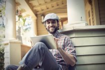 Lächeln Mann mit Mütze mit digitalem Tablet auf Veranda auf dem Land Haus. — Stockfoto