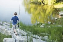 Menino idade elementar andando com vara de pesca na costa do lago no campo . — Fotografia de Stock