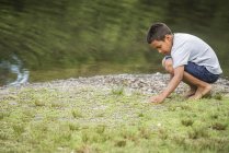 Età elementare ragazzo raccogliendo pietre sulla riva del lago . — Foto stock