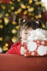 Mädchen im Grundschulalter hält Geschenk mit Schleife vor dem Weihnachtsbaum. — Stockfoto
