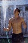 Man in shorts with bare chest holding garden hose and standing in spray of water. — Stock Photo