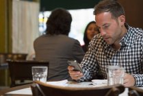 Man at cafe table checking phone with couple talking in background. — Stock Photo