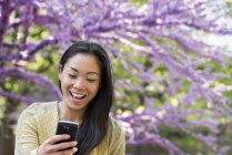Woman looking at phone and laughing in park under tree with pink blossom. — Stock Photo