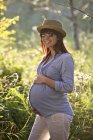 Mujer embarazada en sombrero y gafas de sol con las manos en la barriga en el jardín . - foto de stock