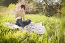 Jeune femme assise dans un champ herbeux sur couverture et en utilisant une tablette numérique . — Photo de stock