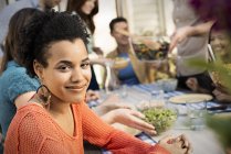 Young woman looking in camera with friends gathering around table and meal. — Stock Photo