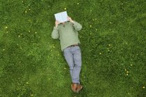 Blick von oben auf einen Mann, der auf dem Rücken auf grünem Gras liegt und Buch liest. — Stockfoto