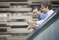 Двое мужчин, опираясь на перила, используя смартфон и улыбаясь на улице . — стоковое фото