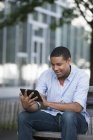 Hombre afroamericano usando tableta digital mientras está sentado en el banco en la ciudad . - foto de stock
