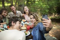 Amici seduti intorno tavolo da picnic e donna prendendo selfie di gruppo . — Foto stock