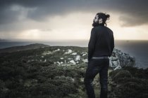 Hombre con barba y pelo moño de pie y mirando sobre el paisaje de montaña al atardecer . - foto de stock