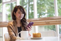 Young woman sitting at table with mug and slice of cake and using smartphone. — Stock Photo