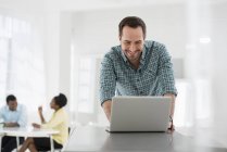 Homem inclinado na mesa e usando laptop com pessoas sentadas e falando no escritório . — Fotografia de Stock